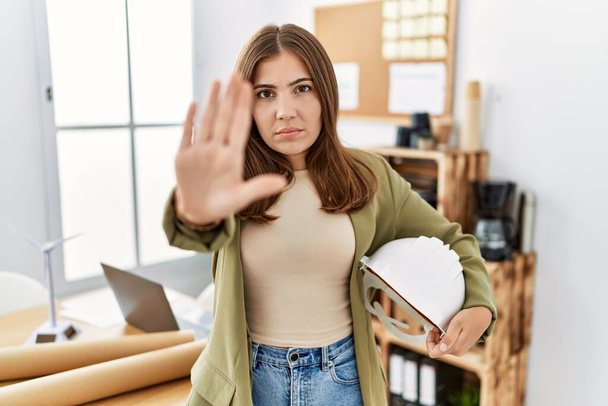 młoda brunetka kobieta trzyma architekt hardhat w biurze z otwartą ręką robi znak stop z poważnym i pewnym siebie wyrazem twarzy, obrona gest  - Zdjęcie, obraz