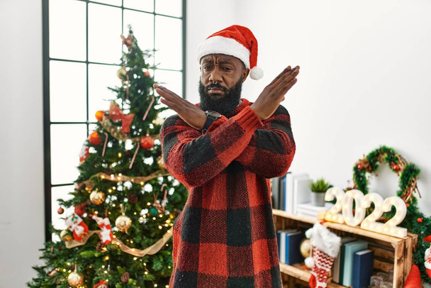 Αφροαμερικάνος που φοράει καπέλο Σάντα Κλάους στέκεται δίπλα στο χριστουγεννιάτικο δέντρο έκφραση απόρριψης διασχίζοντας τα χέρια κάνοντας αρνητικό πρόσημο, θυμωμένο πρόσωπο  - Φωτογραφία, εικόνα