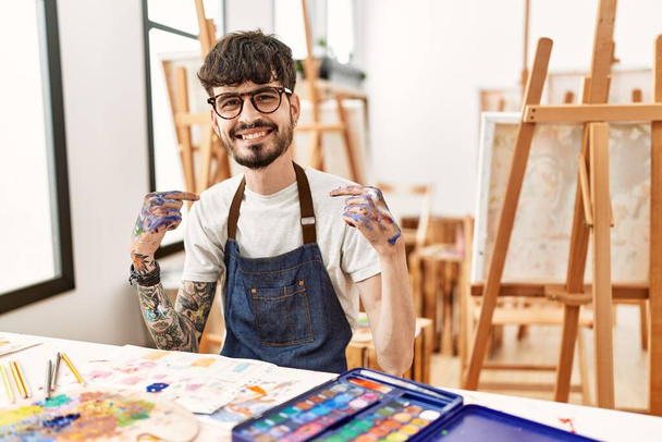 Латиноамериканец с бородой в художественной студии выглядит уверенно с улыбкой на лице, указывая на себя пальцами гордым и счастливым.  - Фото, изображение