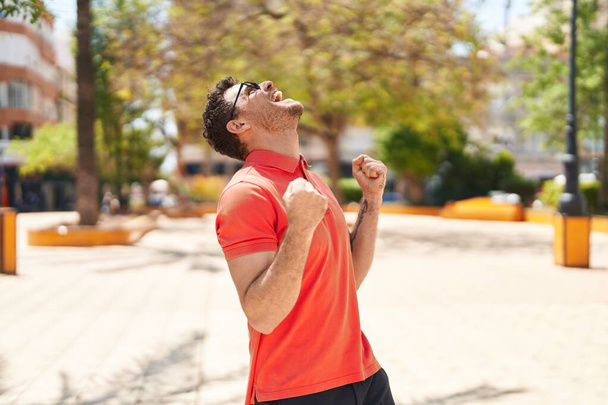 Junger hispanischer Mann mit Brille und feierndem Gesichtsausdruck im Park - Foto, Bild
