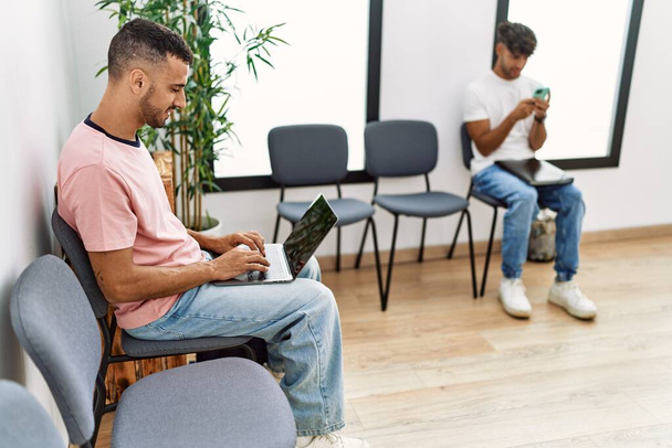 Twee Latijns-Amerikaanse mannen met laptop en smartphone op stoel in de wachtkamer - Foto, afbeelding