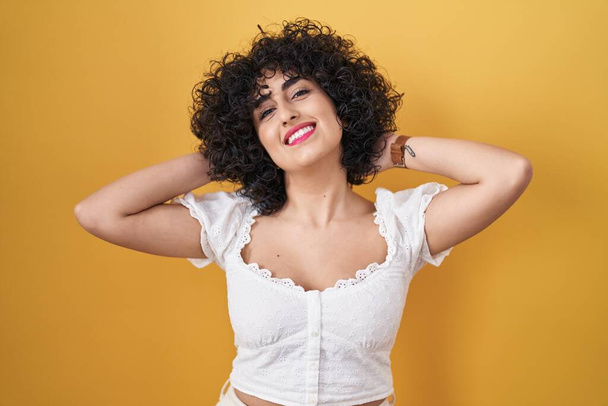 Mladá bruneta žena s kudrnatými vlasy stojící nad žlutým pozadím relaxační a protahování, ruce a ruce za hlavou a krk s úsměvem šťastný  - Fotografie, Obrázek