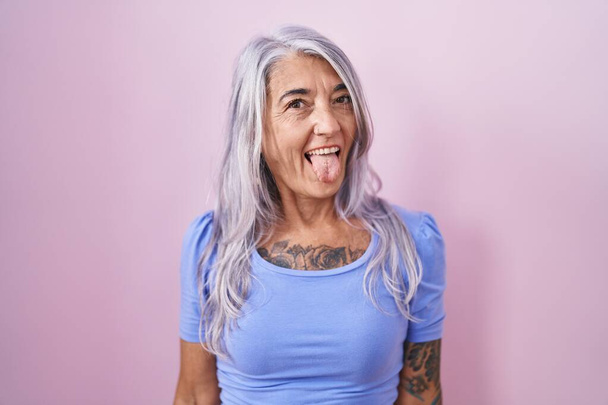 Mujer de mediana edad con tatuajes de pie sobre fondo rosa sacando la lengua feliz con expresión divertida. concepto de emoción.  - Foto, imagen
