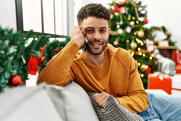 Young arab man talking on the smartphone sitting on the sofa by christmas tree at home. - Photo, Image