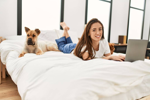 Młoda Latynoska kobieta korzystająca z laptopa leżącego na łóżku z psami w sypialni - Zdjęcie, obraz