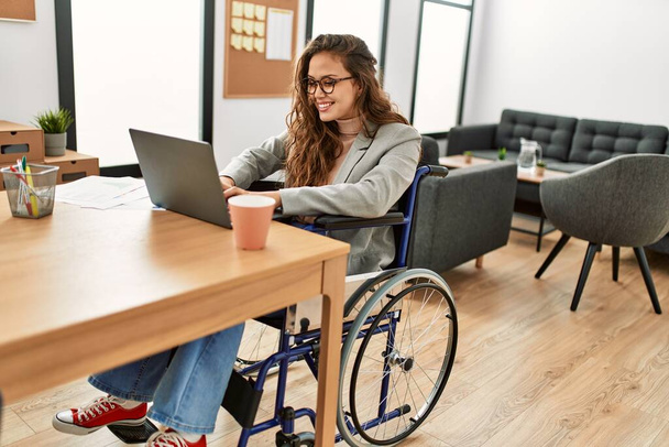 Молодая красивая латиноамериканка бизнес-работница с ноутбуком сидит на инвалидной коляске в офисе - Фото, изображение