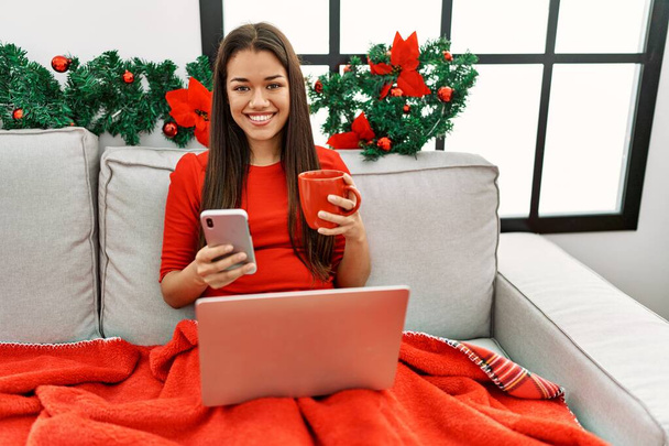 Młoda Latynoska kobieta za pomocą smartfona siedzi na Boże Narodzenie wystrój w domu - Zdjęcie, obraz