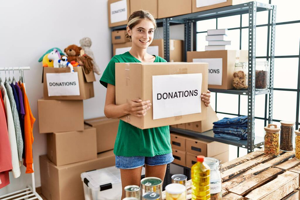 Νεαρή λευκή γυναίκα που φοράει στολή εθελοντή κρατώντας κουτί δωρεών στο φιλανθρωπικό κέντρο - Φωτογραφία, εικόνα