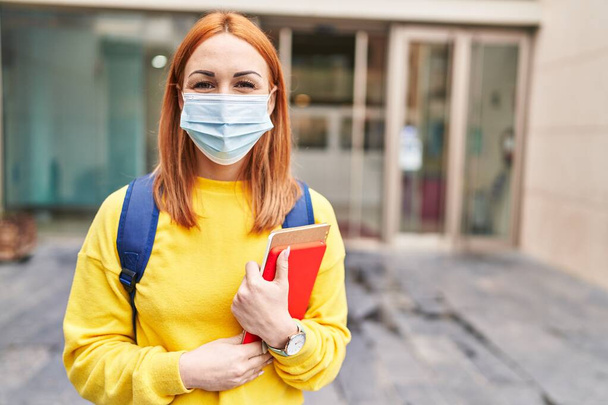 Young woman wearing safety mask and student backpack holding books looking positive and happy standing and smiling with a confident smile showing teeth  - Foto, imagen