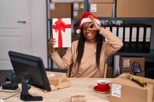 Mladý africký Američan s copánky pracuje v malém podnikání ekommerce na Vánoce s úsměvem šťastný dělá ok znamení s rukou na oku dívá skrz prsty  - Fotografie, Obrázek