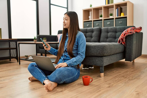 Young chinese girl using laptop and smartphone sitting on the floor at home. - Photo, image