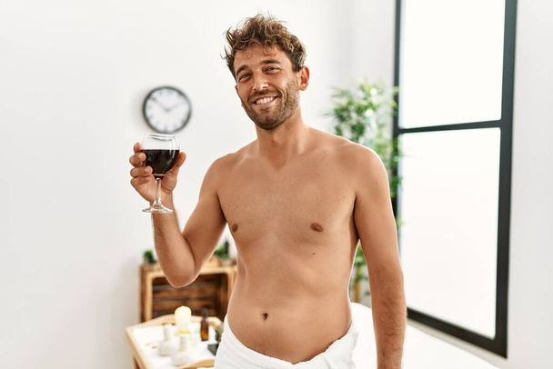 Junger hispanischer Mann lächelt selbstbewusst und trinkt Wein im Schönheitszentrum - Foto, Bild