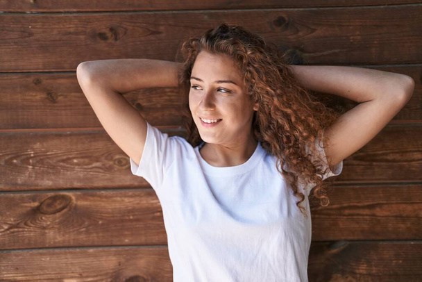 Молодая красивая латиноамериканка улыбается уверенно расслабленной с руками на голове на изолированном деревянном фоне - Фото, изображение