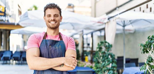 Young hispanic waiter wearing apron standing with arms crossed gesture at coffee shop. - Photo, Image