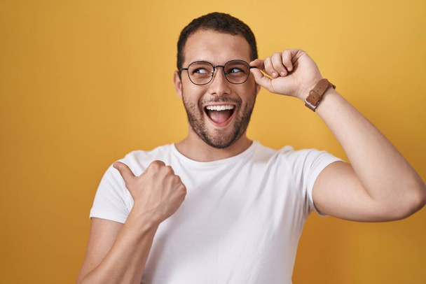 Hispanic man holding glasses pointing thumb up to the side smiling happy with open mouth  - Fotoğraf, Görsel