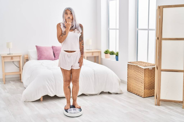 Middle age woman with grey hair standing on weight machine at the bedroom covering mouth with hand, shocked and afraid for mistake. surprised expression  - Photo, Image