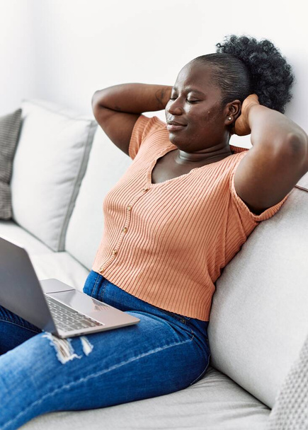 jong afrikaanse amerikaanse vrouw ontspannen met handen op hoofd met behulp van laptop thuis - Foto, afbeelding