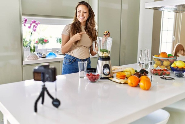 Joven hermosa mujer hispana preparando batido de verduras con licuadora grabación video tutorial con cámara en la cocina - Foto, Imagen