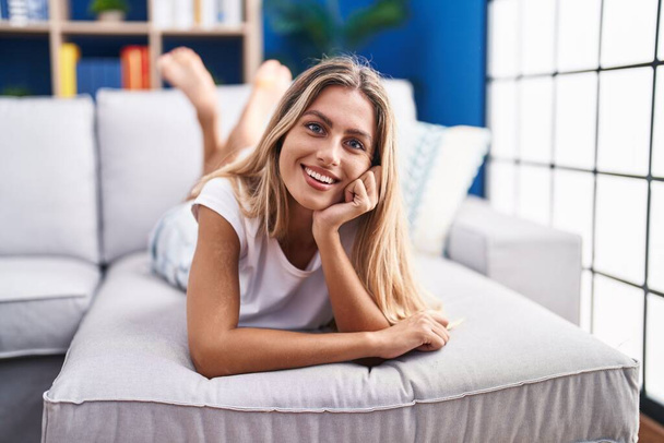 Молодая блондинка, уверенно улыбающаяся, лежа дома на диване - Фото, изображение
