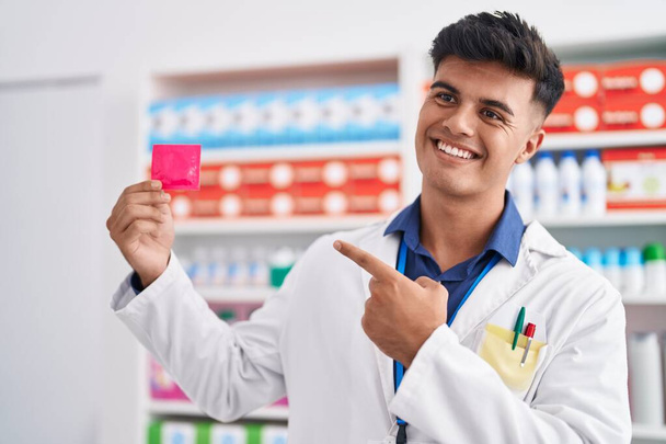 Jovem hispânico homem farmacêutico sorrindo confiante segurando preservativo na farmácia - Foto, Imagem
