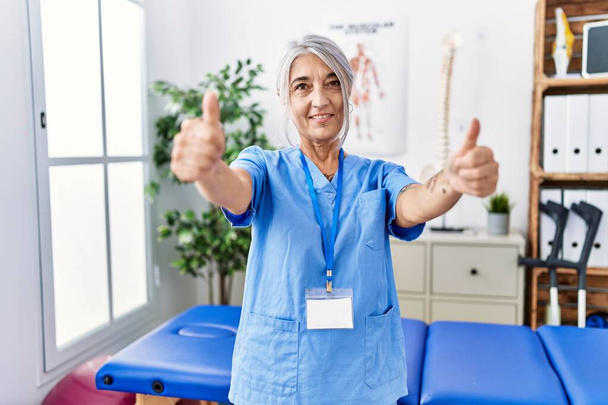 Middle age grey-haired woman wearing physiotherapist uniform at medical clinic approving doing positive gesture with hand, thumbs up smiling and happy for success. winner gesture.  - Photo, Image