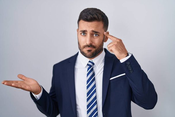 Hombre hispano guapo vistiendo traje y corbata confundido y molesto con la palma abierta mostrando espacio de copia y señalando con el dedo a la frente. Piénsalo.  - Foto, Imagen