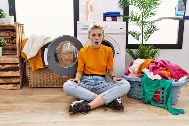 Junge blonde Frau beim Wäschewaschen sitzt ängstlich an der Waschmaschine und schockiert mit Überraschung und erstauntem Gesichtsausdruck, Angst und aufgeregtem Gesicht.  - Foto, Bild
