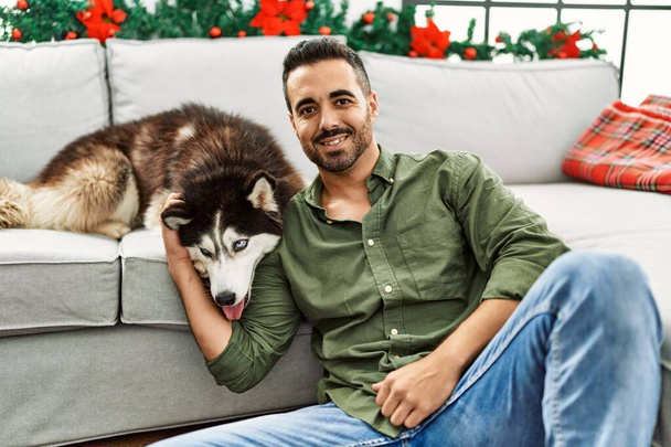 Evdeki Noel dekorunda köpekle kanepede otururken kendinden emin gülümseyen İspanyol genç adam. - Fotoğraf, Görsel