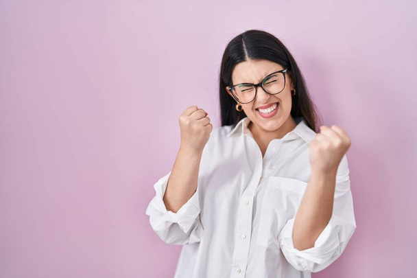Young brunette woman standing over pink background very happy and excited doing winner gesture with arms raised, smiling and screaming for success. celebration concept.  - Фото, изображение