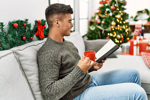 Young hispanic man reading book drinking coffee sitting on sofa by christmas tree at home - 写真・画像