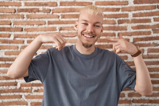 Homem caucasiano jovem em pé sobre a parede de tijolos sorrindo alegre mostrando e apontando com os dedos dentes e boca. conceito de saúde dentária.  - Foto, Imagem