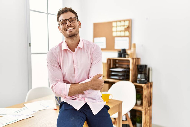 Jonge Spaanse man lacht vol vertrouwen met armen gekruist gebaar op kantoor - Foto, afbeelding
