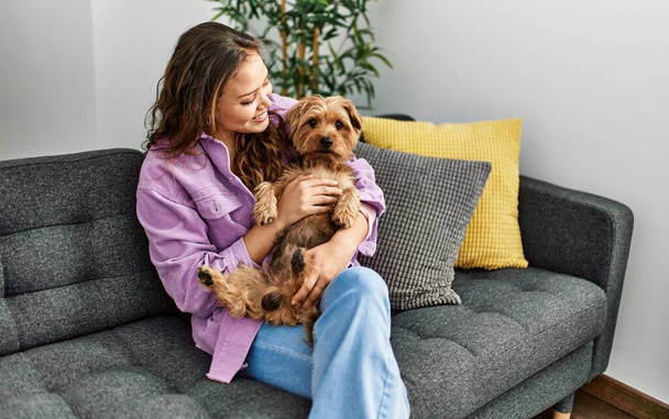 Νέα όμορφη ισπανόφωνη γυναίκα αγκαλιάζει το σκυλί κάθεται στον καναπέ στο σπίτι - Φωτογραφία, εικόνα