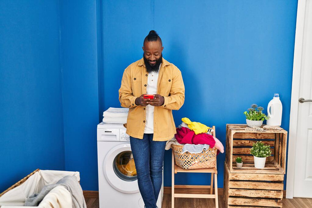 Jonge Afrikaans-Amerikaanse man met behulp van smartphone wachten op wasmachine in de wasruimte - Foto, afbeelding