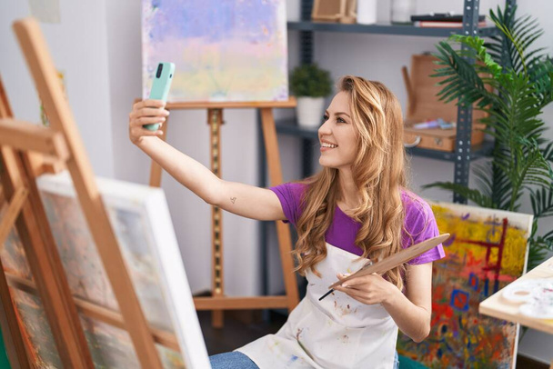 Νεαρή ξανθιά γυναίκα καλλιτέχνης χαμογελώντας αυτοπεποίθηση κάνουν selfie από το smartphone στο στούντιο τέχνης - Φωτογραφία, εικόνα