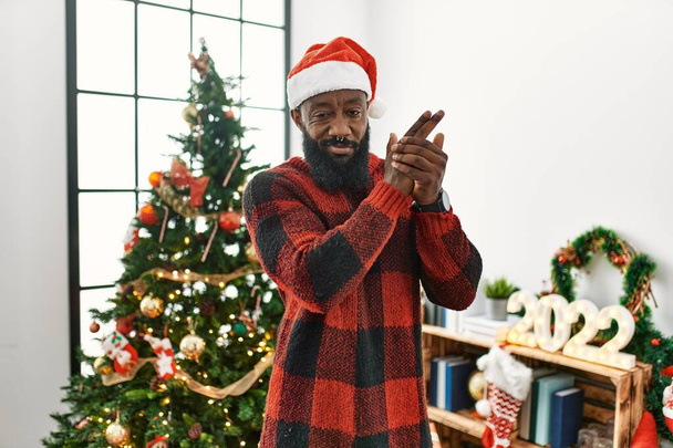 Hombre afroamericano con sombrero de Santa Claus de pie junto al árbol de navidad sosteniendo un arma simbólica con gesto de mano, jugando a matar armas de fuego, cara enojada  - Foto, imagen