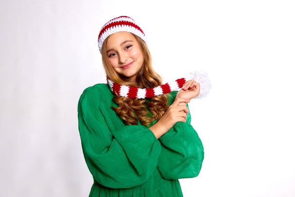 fille heureuse en robe verte de Noël et chapeau tricoté rayé rouge regardant la caméra sur fond blanc - Photo, image