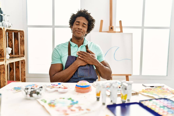 若いアフリカ系アメリカ人の男性がアートスタジオのテーブルの上に座って、目を閉じて胸に手を笑顔で、顔に感謝のジェスチャー。健康の概念.  - 写真・画像