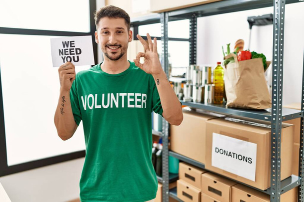 Joven hombre hispano con camiseta voluntaria sosteniendo necesitamos que usted banner haciendo signo bien con los dedos, sonriendo gesto amistoso excelente símbolo  - Foto, Imagen