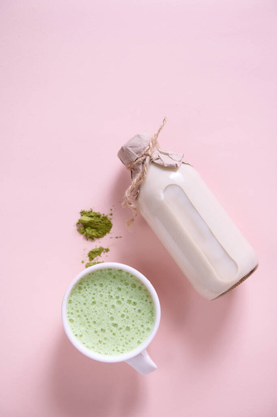 Flat lay. Vertical studio shot of a white ceramic cup of whipped matcha latte with foam, heap of powdered green tea, bottle of healthy organic wholesome raw vegan milk over pink background. Copy space - Фото, изображение