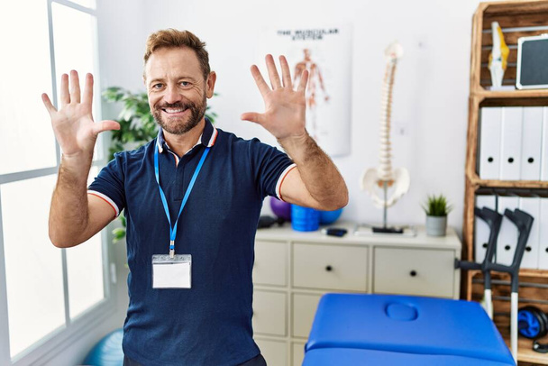 Hombre fisioterapeuta de mediana edad que trabaja en la clínica de recuperación del dolor mostrando y señalando con los dedos el número diez mientras sonríe confiado y feliz.  - Foto, imagen