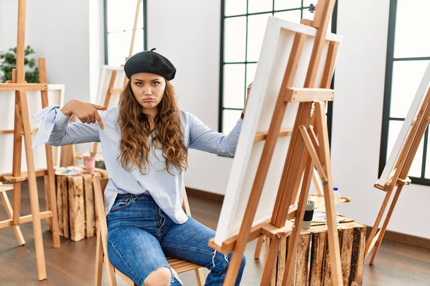 Młoda latynoska artystka malująca na płótnie w pracowni sztuki, patrząc smutno i smutno, wskazując kierunek palcami, nieszczęśliwie i przygnębiona.  - Zdjęcie, obraz
