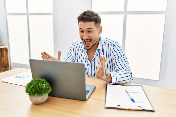 Ofiste çalışan sakallı yakışıklı genç adam bilgisayarını kullanarak kollarını kaldırarak ve açık gözlerle başarıyı kutluyor. Kazanan konsept.  - Fotoğraf, Görsel