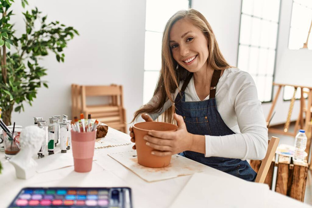 Молода біла жінка посміхається впевнено робить глиняний горщик ручної роботи в художній студії
 - Фото, зображення