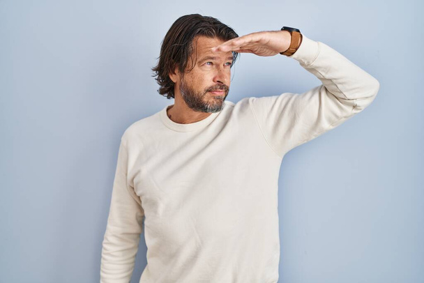 Handsome middle age man wearing casual sweater over blue background very happy and smiling looking far away with hand over head. searching concept.  - Foto, Bild