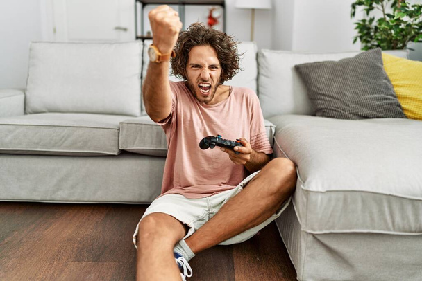 Молодой латиноамериканец, играющий в видеоигру с контроллером дома, раздраженный и разочарованный, кричащий от гнева, кричащий сумасшедший от злости и поднятый рукой  - Фото, изображение