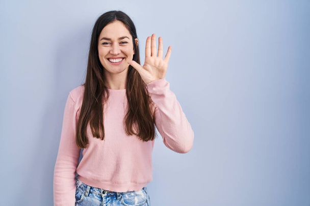 Giovane donna bruna in piedi sopra lo sfondo blu mostrando e indicando con le dita il numero cinque sorridendo fiducioso e felice.  - Foto, immagini