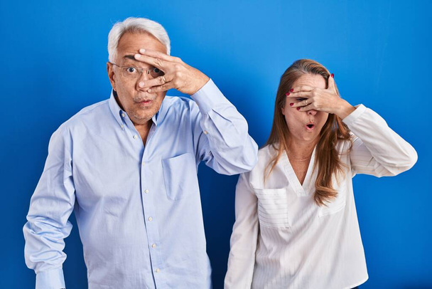 Couple hispanique du moyen âge debout sur fond bleu regardant sous le choc couvrant le visage et les yeux avec la main, regardant à travers les doigts avec une expression embarrassée.  - Photo, image