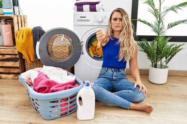 Young beautiful woman doing laundry sitting by wicker basket looking unhappy and angry showing rejection and negative with thumbs down gesture. bad expression.  - Foto, Imagem