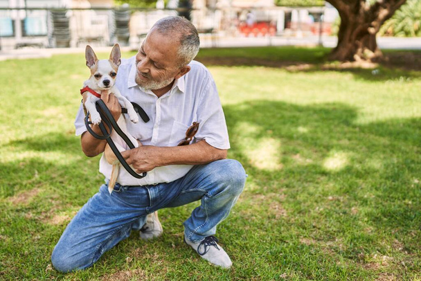Senior man glimlachend zelfverzekerde knuffelende chihuahua in park - Foto, afbeelding